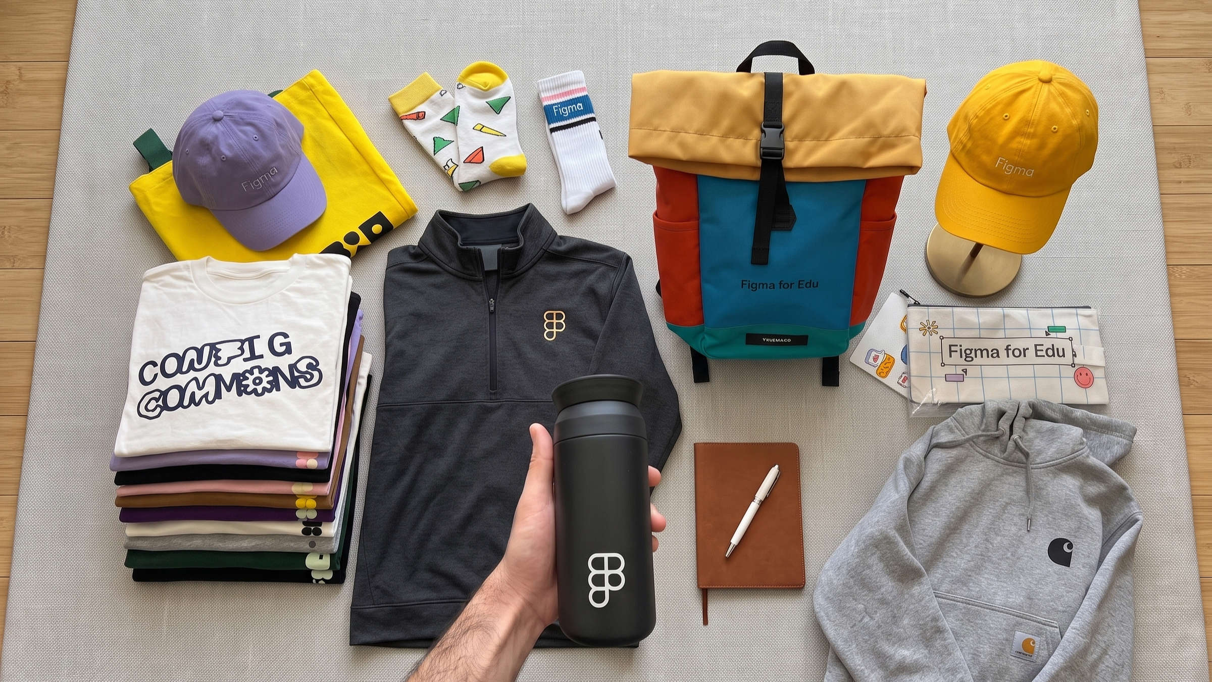Overhead flat-lay of a welcome kit with stacked tees, a quarter-zip, hoodie, two caps, backpack, tumbler, and notebook