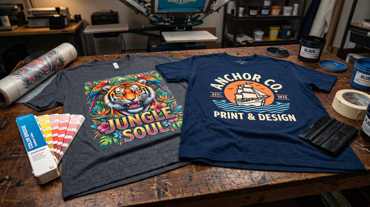 Full-color tiger print tee beside a two-color logo tee on a print shop bench with a Pantone swatch book