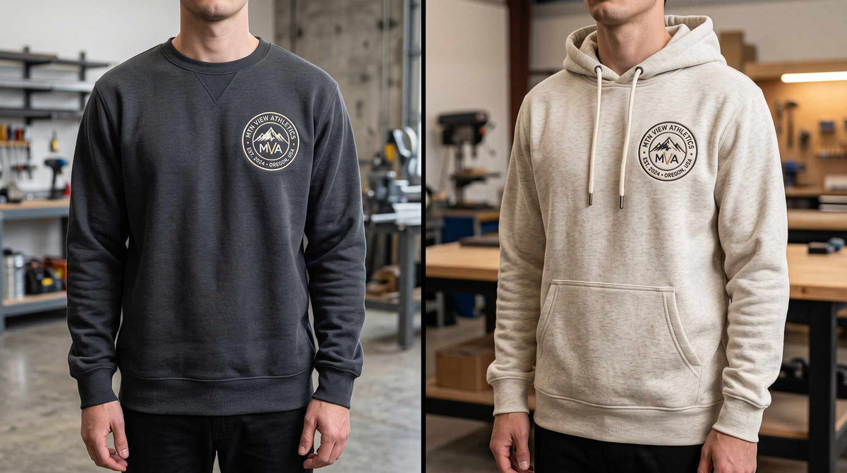 The same left-chest logo on a charcoal crewneck sweatshirt and a heather cream hoodie, worn side by side in a workshop