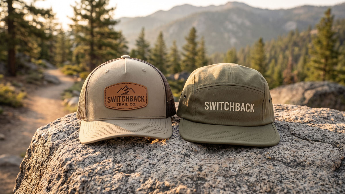 Five-panel trucker with a SWITCHBACK TRAIL CO. leather patch beside an olive camp cap on a boulder at a trailhead